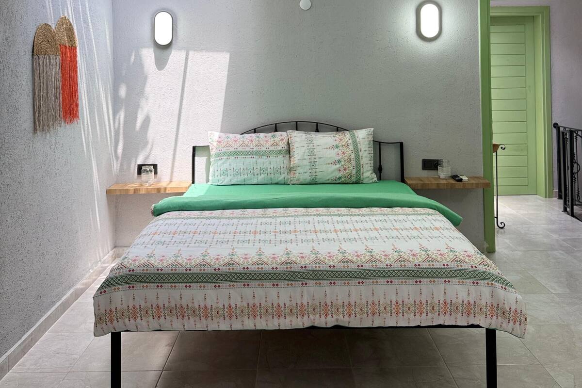 Quarto com cama, mesa de cabeceira, porta verde e luminária fixada na parede.