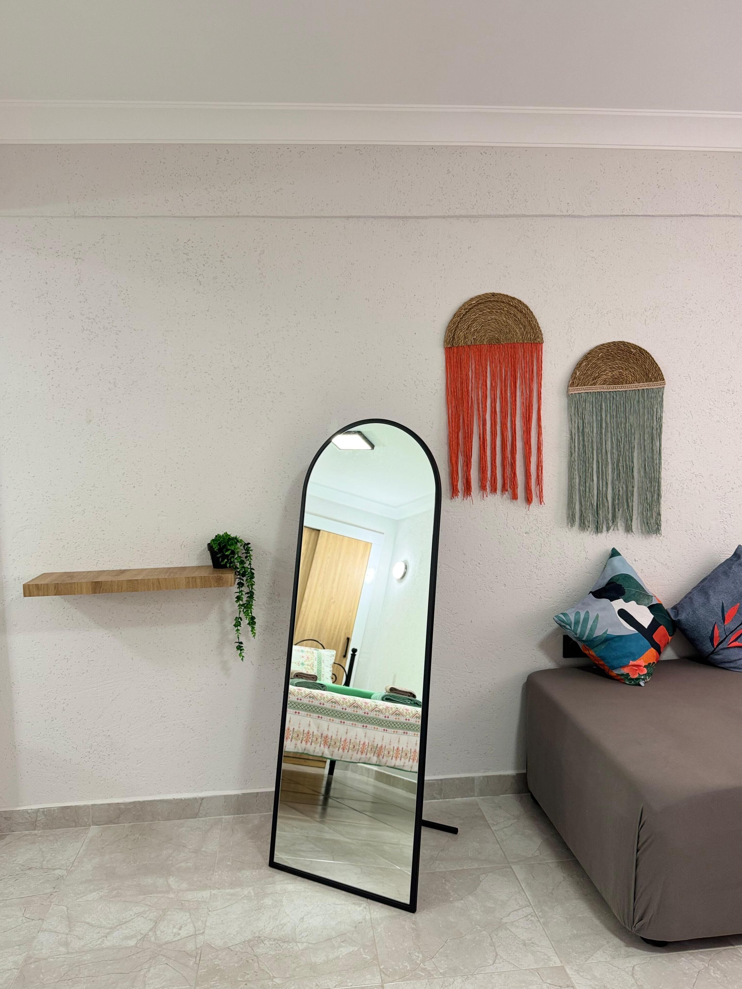 A room with a large mirror, a wooden shelf with a plant, a couch with patterned cushions, and two wall-mounted decorative items.