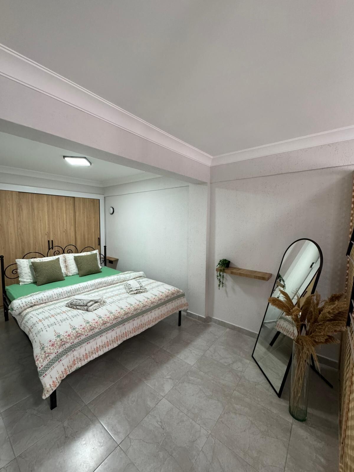A bedroom with a bed, a nightstand, a mirror, and a shelf with a plant.