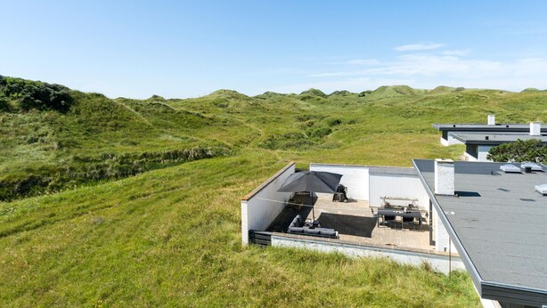 Een modern huis met een plat dak, een terras met tuinmeubilair en uitzicht op glooiende heuvels.