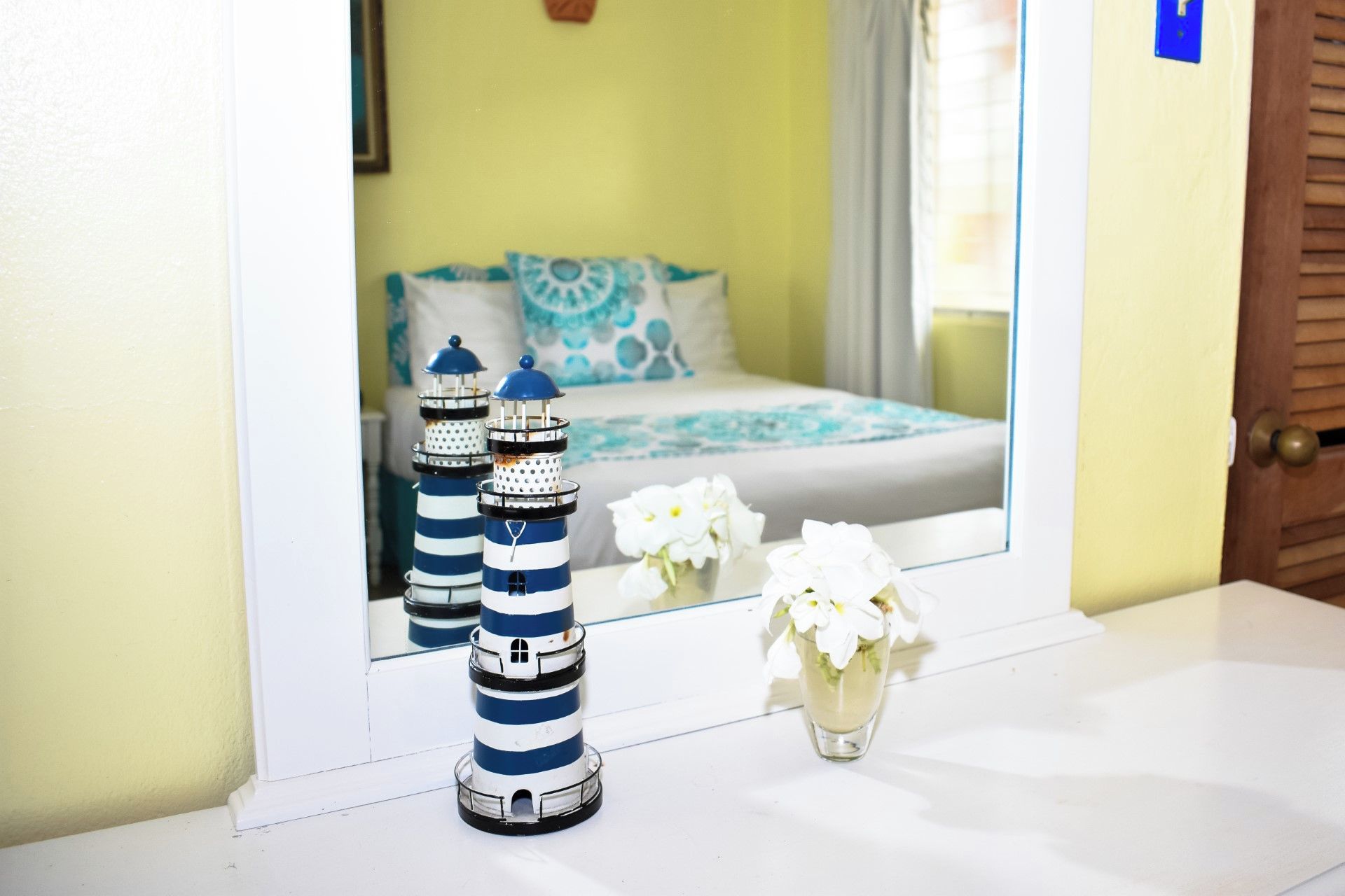 A bathroom with a mirror reflecting a bed with blue and white bedding, a white vase with white flowers, and two striped lighthouses on the counter.