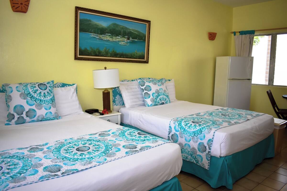 Quarto de hotel com duas camas, um quadro de uma paisagem costeira, uma mesa de cabeceira com uma luminária e uma geladeira.