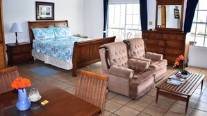 Habitación de hotel con una cama, dos sillones, una mesa de madera, una mesita, una lámpara, un florero con flores y una ventana con cortinas.