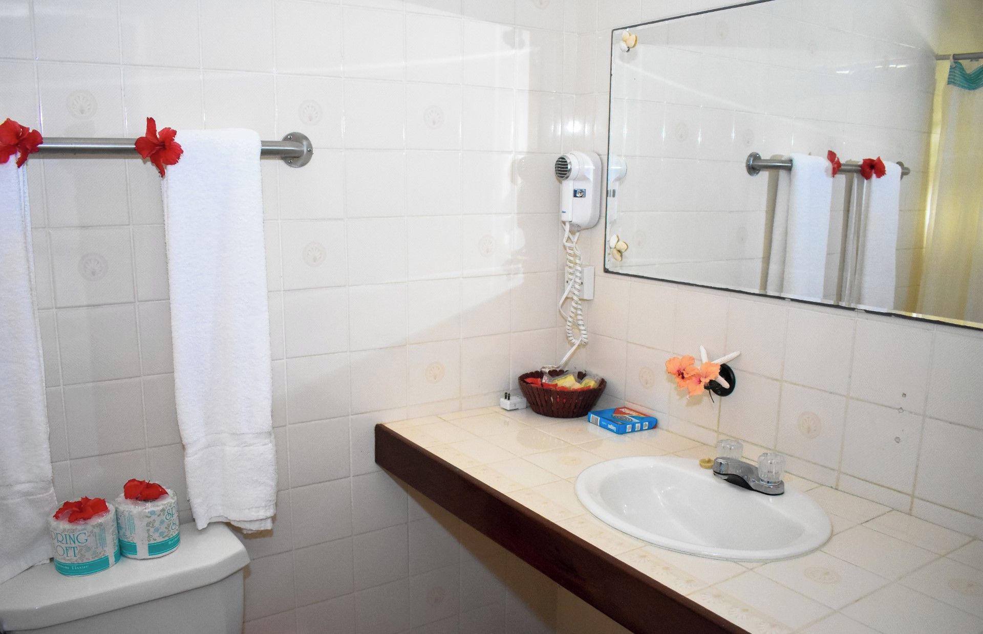 Ein Badezimmer mit weißen Fliesen, einem Waschbecken, einem Spiegel, einem Fön und einem Korb mit Toilettenartikeln.