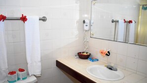 Un baño con azulejos blancos, un lavamanos, un espejo, un secador de pelo y una cesta con artículos de aseo.