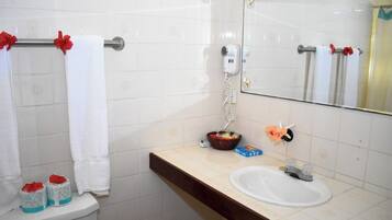 Ein Badezimmer mit weißen Fliesen, einem Waschbecken, einem Spiegel, einem Fön und einem Korb mit Toilettenartikeln.