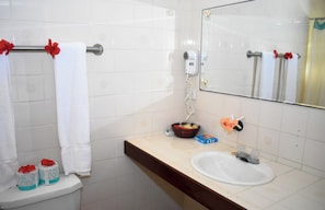 A bathroom with white tiles, a sink, a mirror, a hairdryer, and a basket with toiletries. - The Catamaran Hotel (Falmouth Harbour)