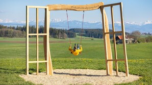 Un columpio de madera con un letrero de hashtag, frente a un campo verde y montañas a lo lejos.