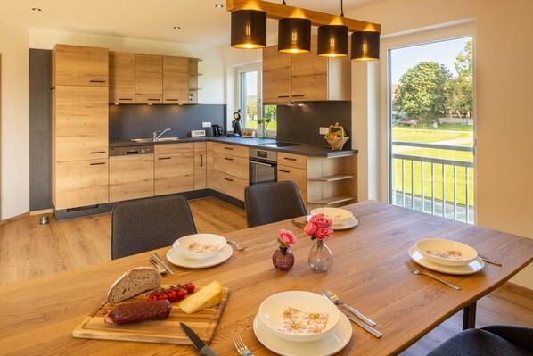 A modern kitchen with wooden cabinets, a dining table set with plates, a vase with flowers, and a view of a green landscape.