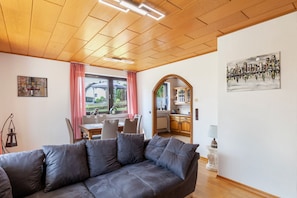 A modern living room with a wooden ceiling, a grey sofa, a dining area with chairs, a kitchen with wooden cabinets, and a painting on the wall. - Apartment 'Rosi's Ferienwohnung' with Mountain View, Shared Terrace and Wi-Fi (Daun)