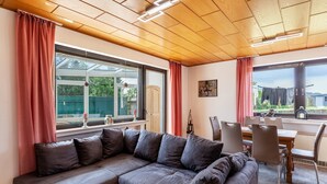 A modern living room with a sectional sofa, dining table, and chairs, large windows with curtains, and a wooden ceiling. - Rosi's Apartment with Mountain View, Shared Terrace and Wi-Fi (Daun)