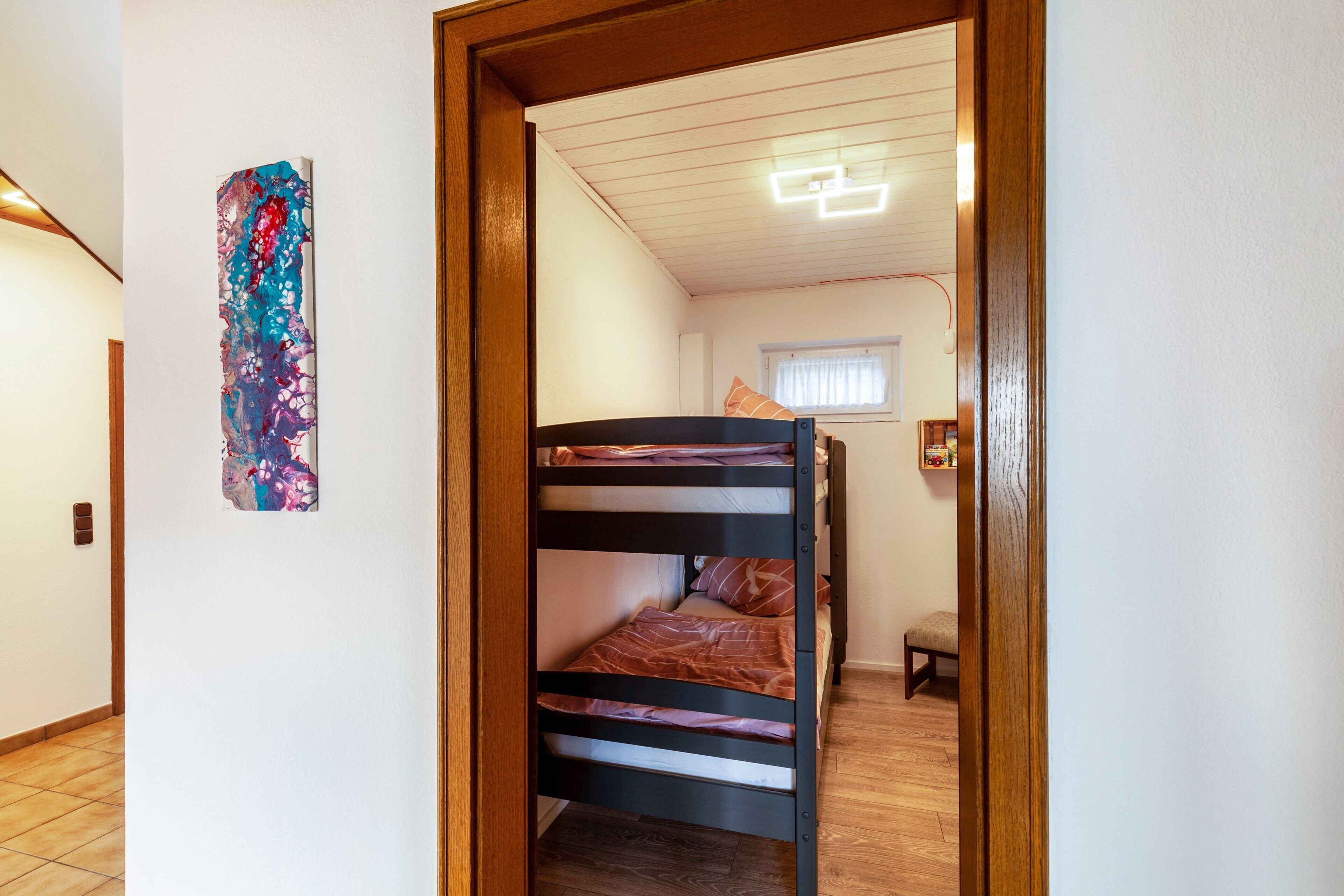 Una cama litera en una habitación con suelo de madera, una ventana con cortinas y una obra de arte abstracta colgada en la pared.
