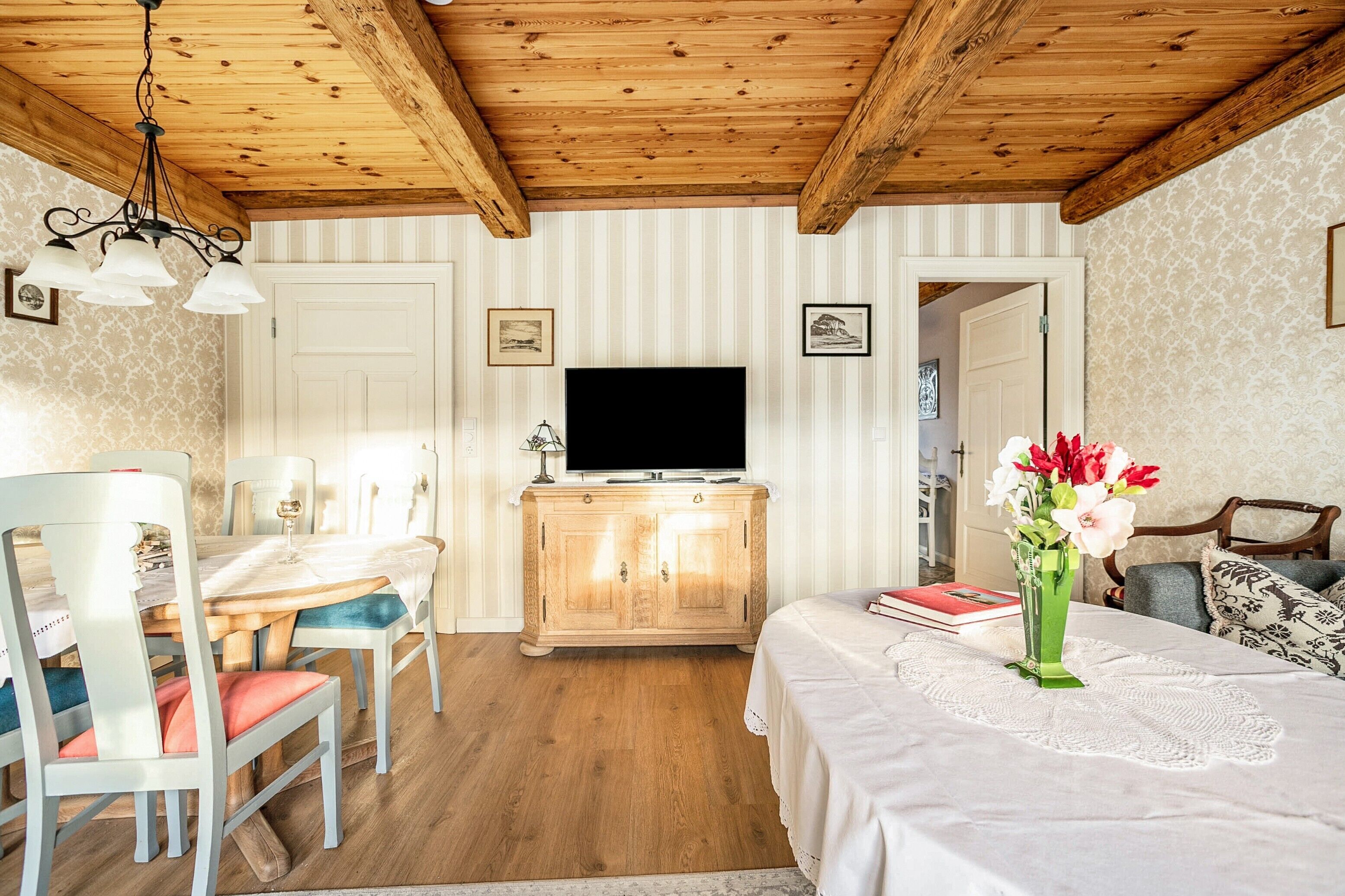 A room with a wooden ceiling, a television on a cabinet, a dining table with chairs, and a bed with a white bedspread.