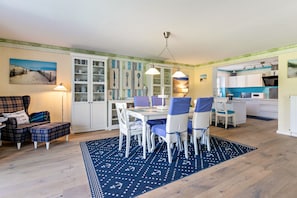 Un comedor con una mesa preparada para cuatro personas, rodeada de sillas con cojines azules. A la izquierda, un armario blanco con puertas de vidrio, y al fondo se ve una cocina.