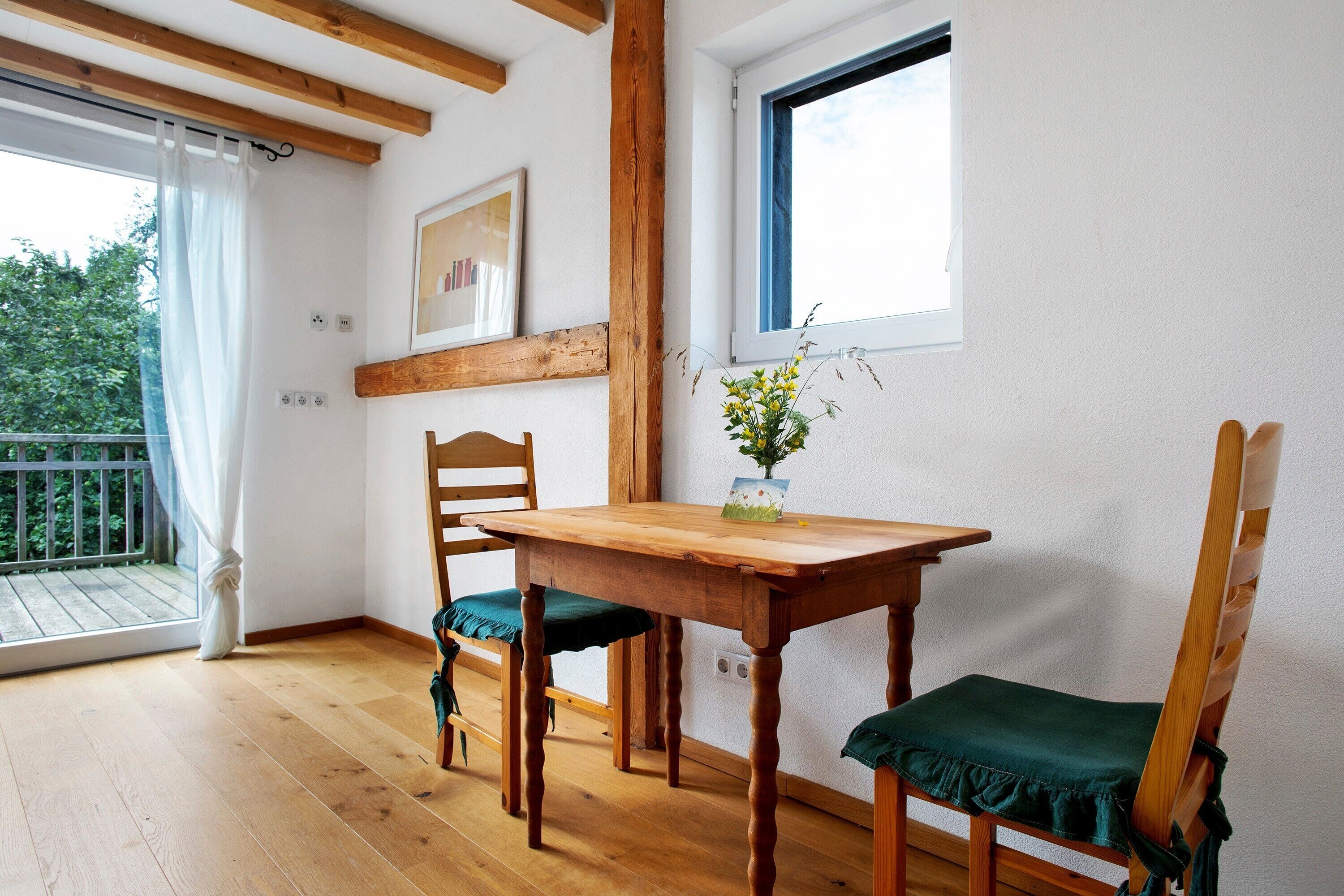 Une table de salle à manger en bois, accompagnée de deux chaises, une fenêtre donnant sur un beau paysage et un balcon avec un rideau.