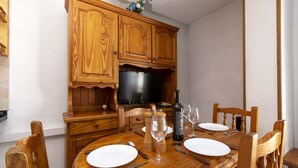 A wooden dining table set with plates, cutlery, and wine glasses, with a television and wooden cabinets in the background. - Las Floritas 4 (Playa de Las Americas)