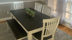 A dining area with a dark wooden table, white chairs, a chandelier, a painting, and a window with curtains.