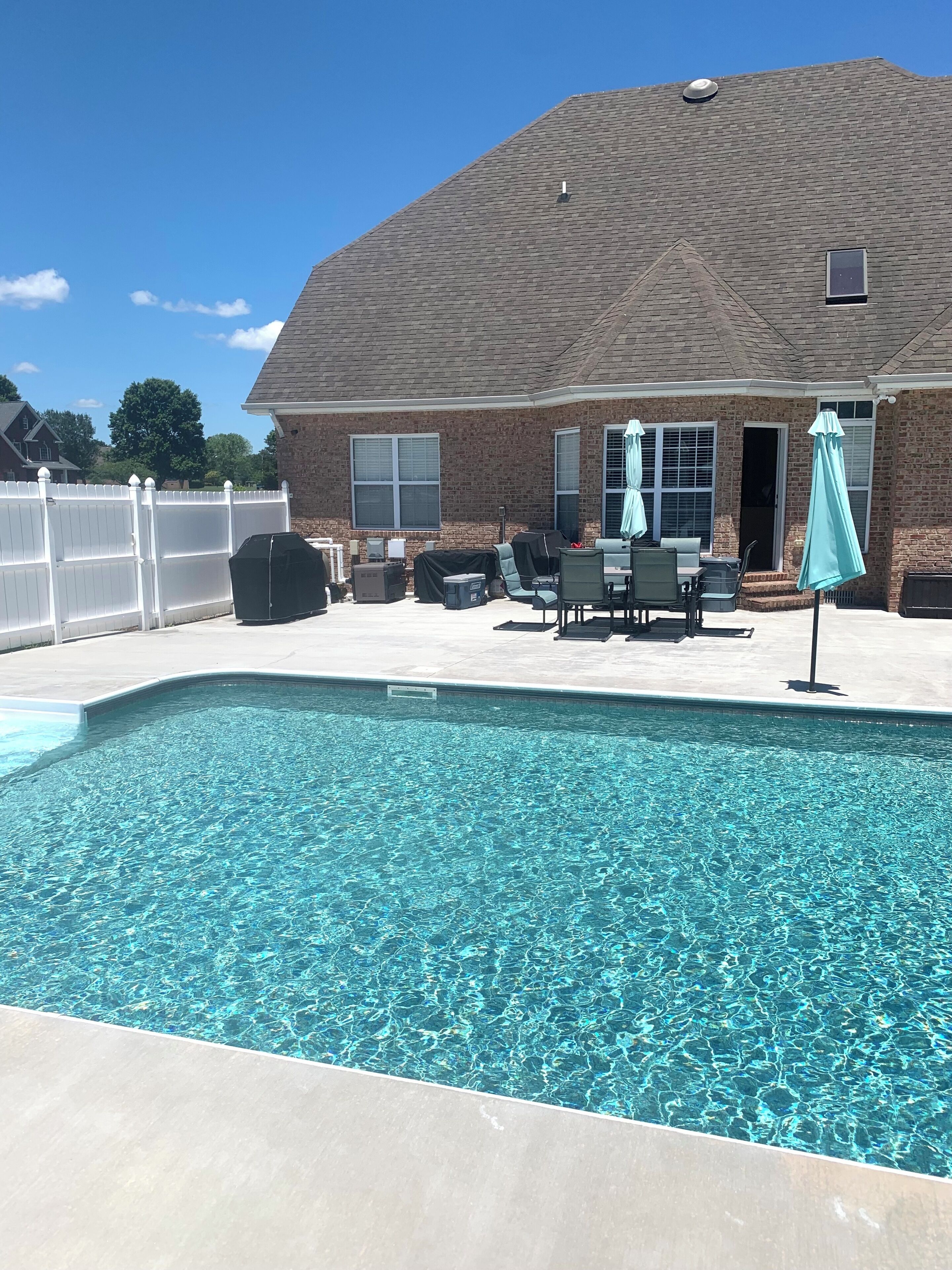 En swimmingpool med fliseterrasse, en overdækket patio med udendørs møbler, og en murstensbygning i baggrunden.