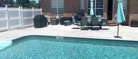 En swimmingpool med fliseterrasse, en overdækket patio med udendørs møbler, og en murstensbygning i baggrunden.