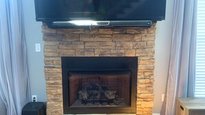 A living room with a stone fireplace, a flat-screen TV mounted above it, and a wooden floor.