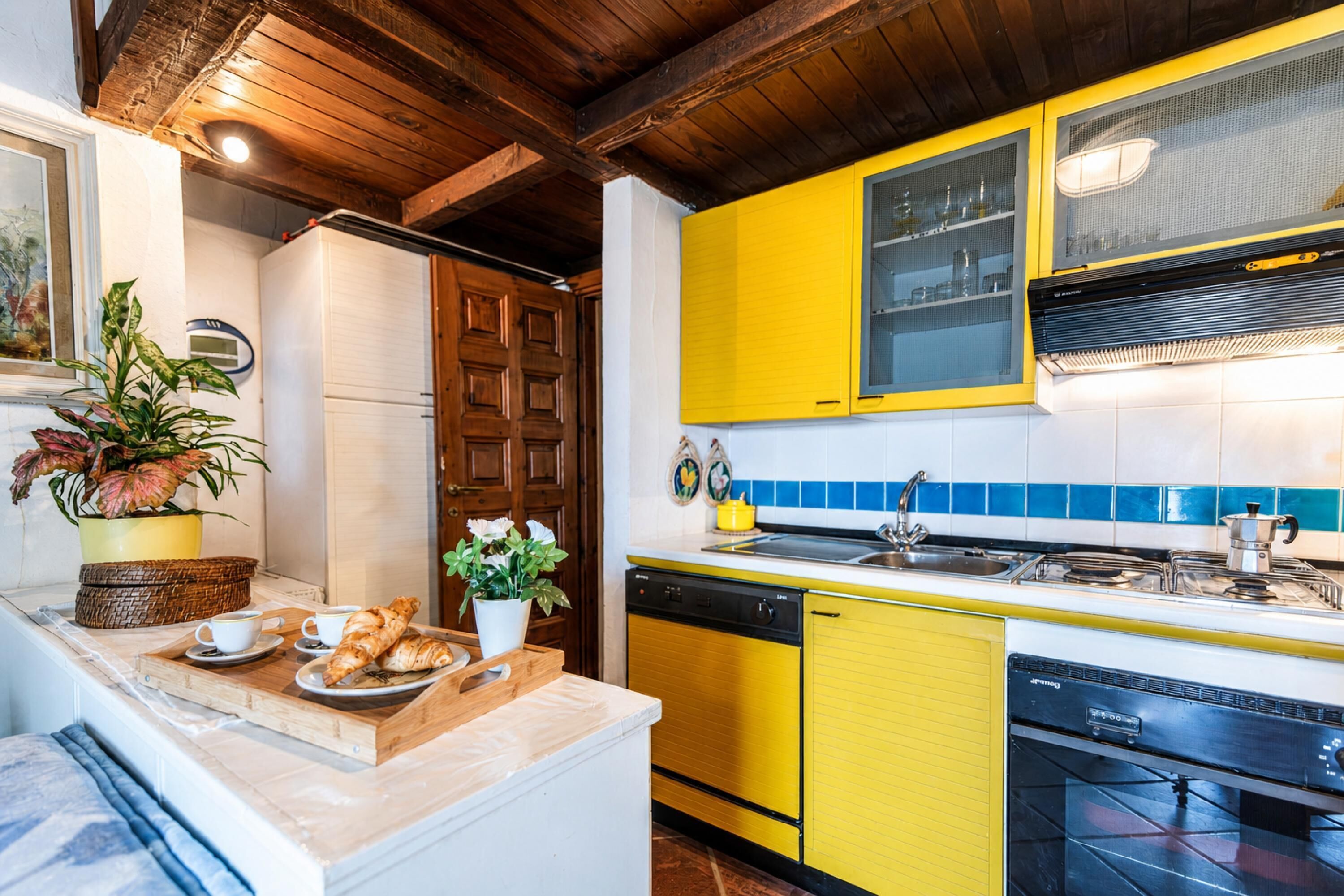 Een keuken met gele kastjes, een witte koelkast en een houten plafond.