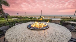 Ein runder Gartentisch mit Feuerstelle in der Mitte, umgeben von Korbstühlen, vor einer Strandkulisse und einem Sonnenuntergangshimmel.