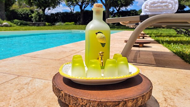 En vandflaske med citronvand og korkprop, omgivet af seks glas på en træbakke, placeret på en terrasse med en swimmingpool i baggrunden.