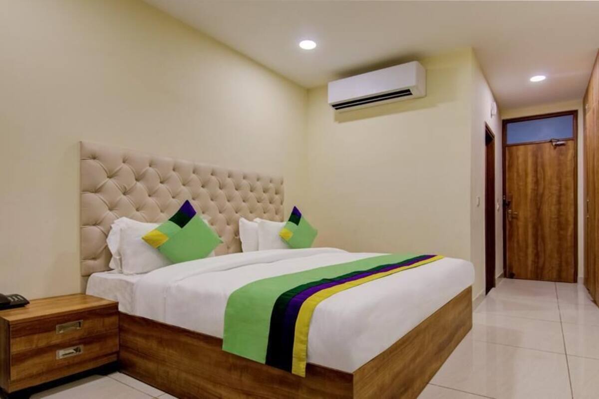 Une chambre à coucher avec une tête de lit capitonnée, un lit avec une literie blanche et des coussins colorés, une table de chevet avec un téléphone et un appareil de climatisation fixé au mur.