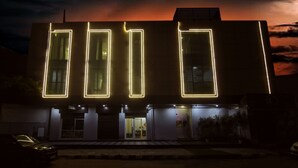 Un edificio con contornos rectangulares iluminados en la fachada, una entrada visible y un coche estacionado al frente.