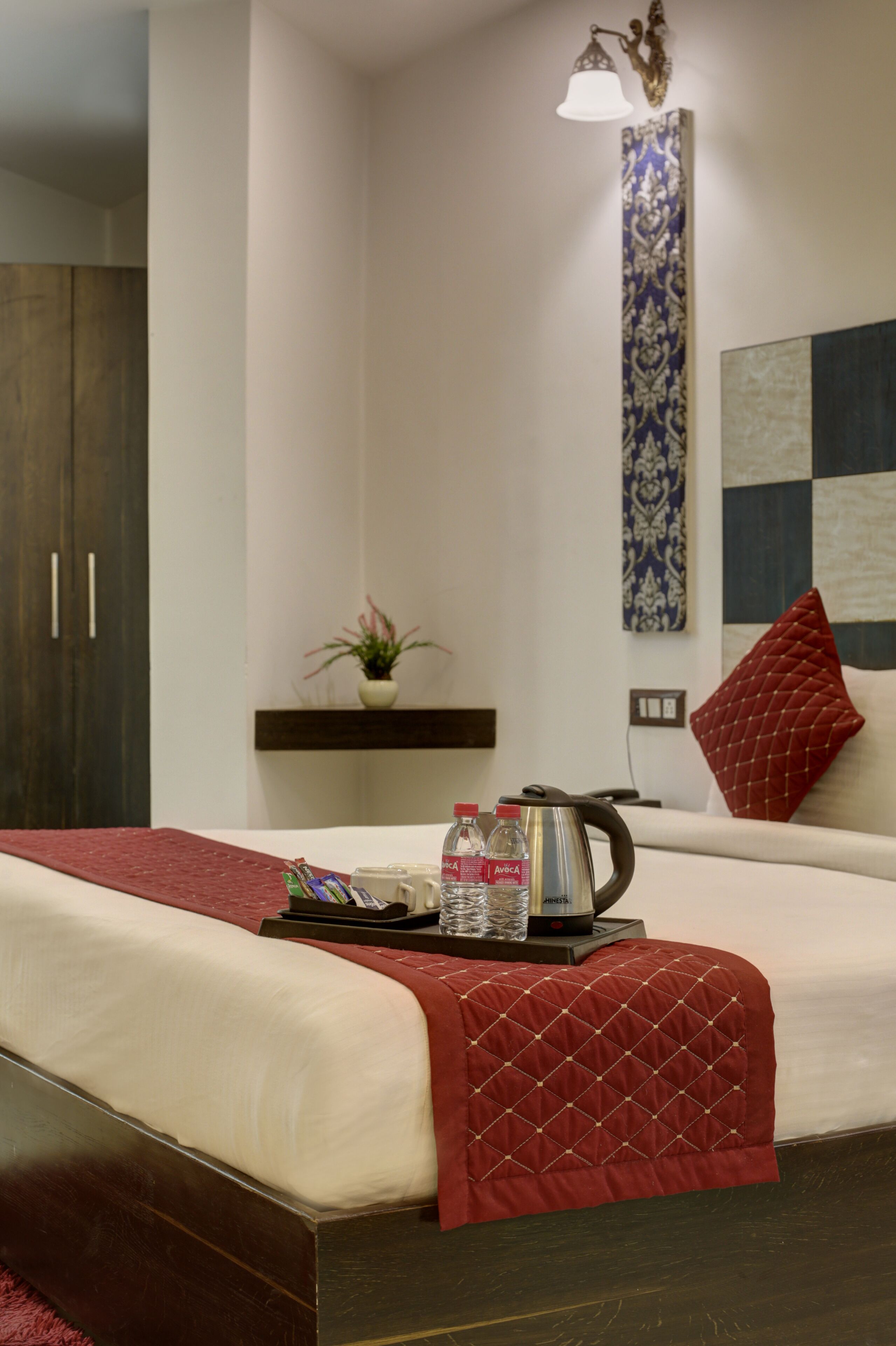 A neatly made bed with a red quilt, a bedside table with a kettle, water bottles, and a tray, a red pillow, a wall-mounted shelf with a plant, and a decorative light fixture.