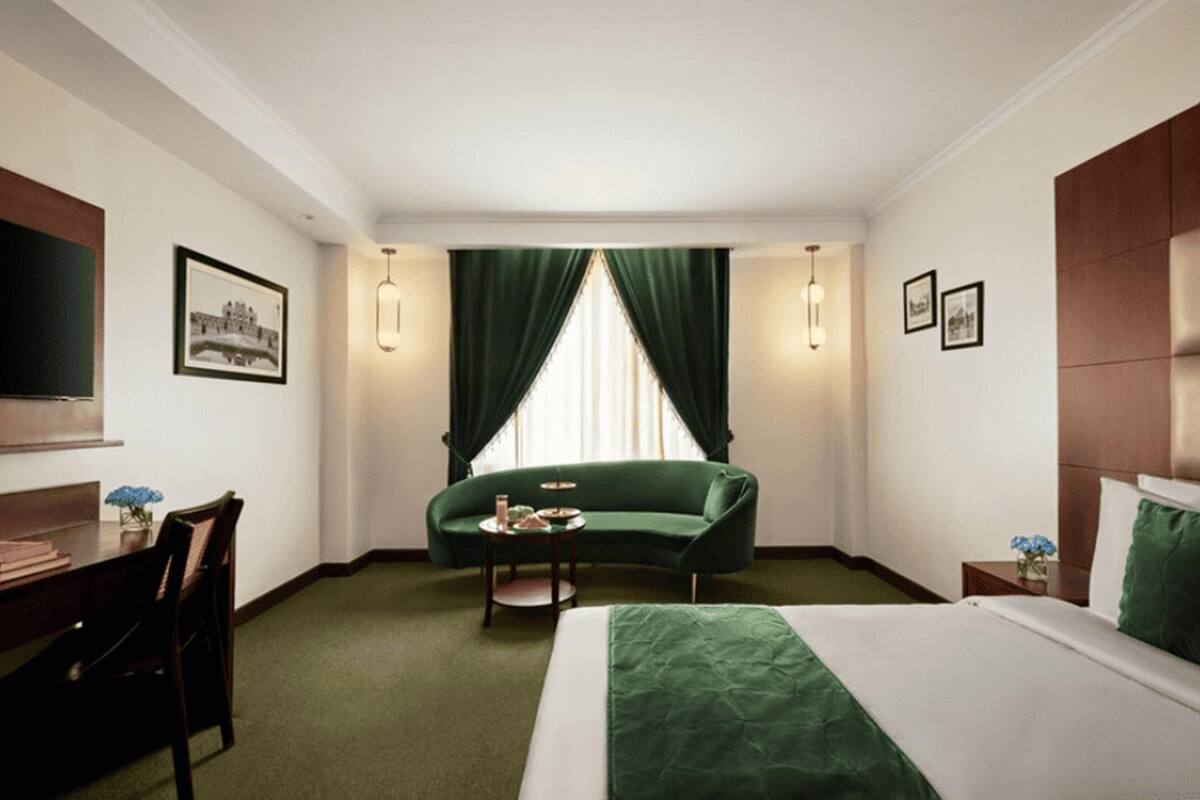 Quarto de hotel com cama, sofá verde, mesa pequena, escrivaninha e televisão.