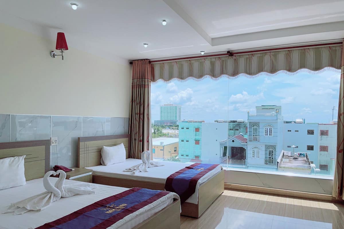 Habitación de hotel con dos camas, un ventanal amplio con vista a la ciudad, una lámpara de pared roja e iluminación en el techo.