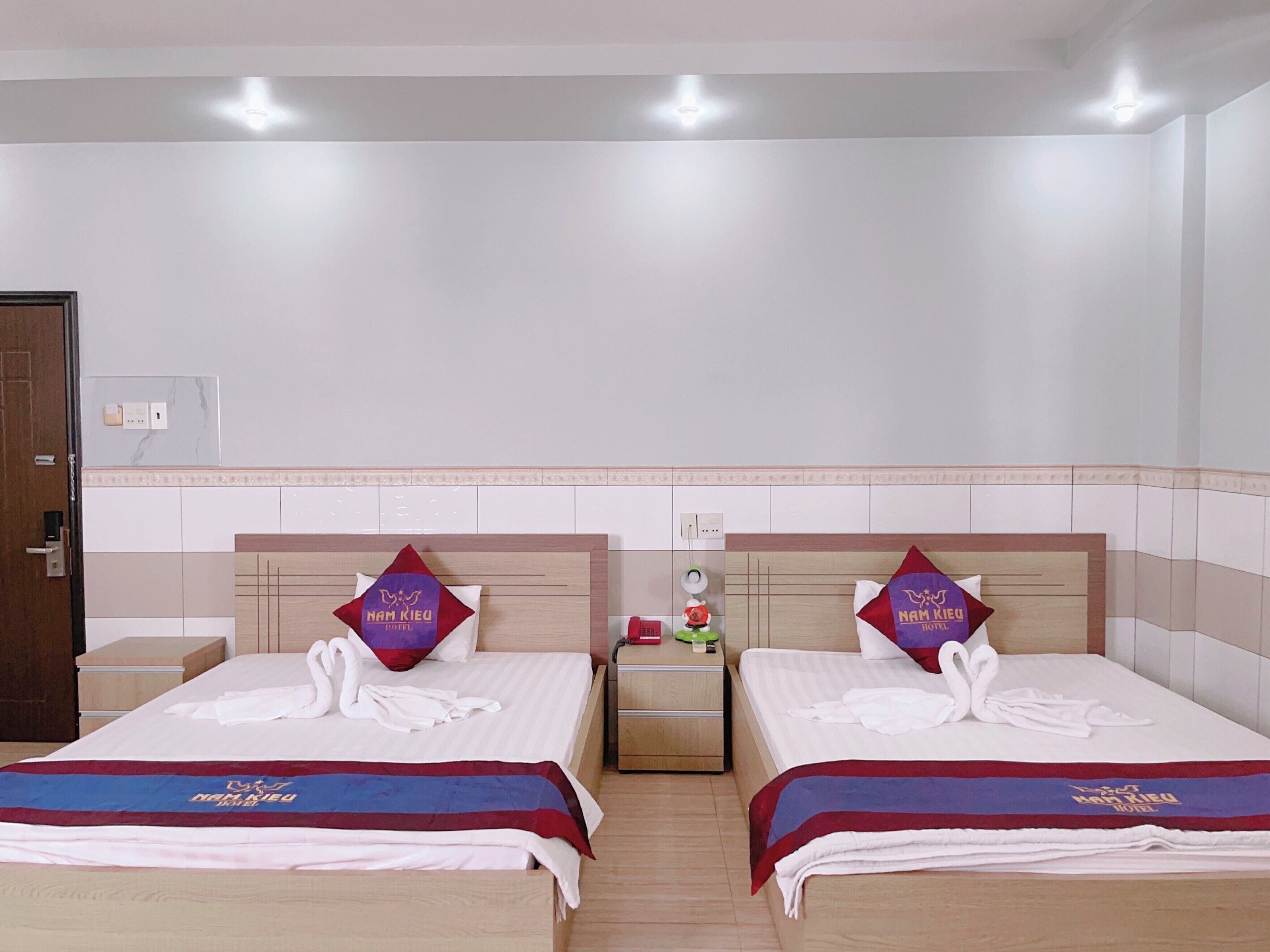 Habitación de hotel con dos camas, cada una con una toalla con forma de cisne y una almohada roja con el logotipo del hotel. Hay mesitas de noche con lámparas y una pequeña mesa con un florero.