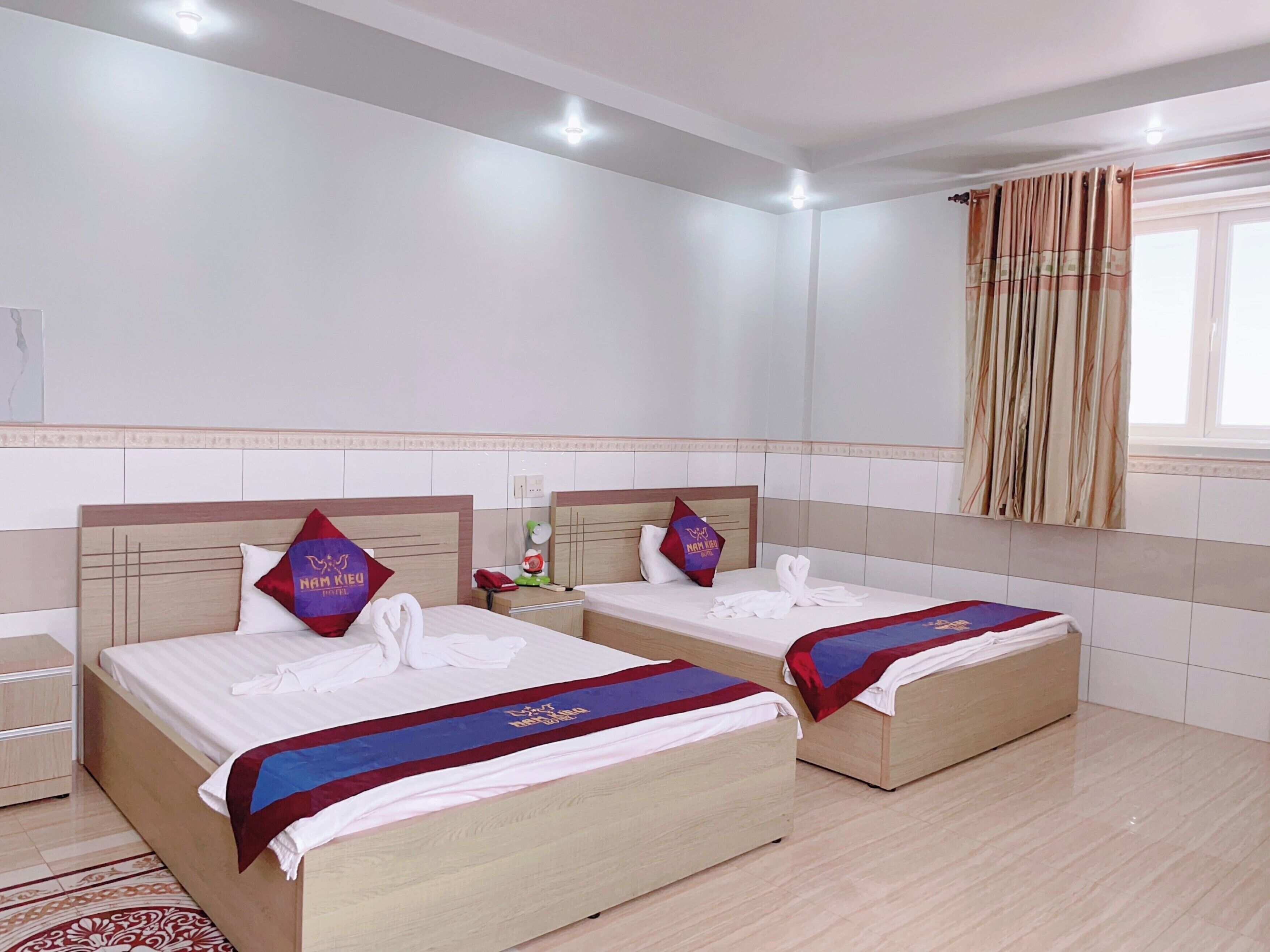 Ein Hotelzimmer mit zwei Betten, jeweils mit Kopfstütze, Nachttisch und Telefon. Das Zimmer verfügt über ein Fenster mit Vorhängen, eine Wand mit Fliesen und eine Deckenleuchte.