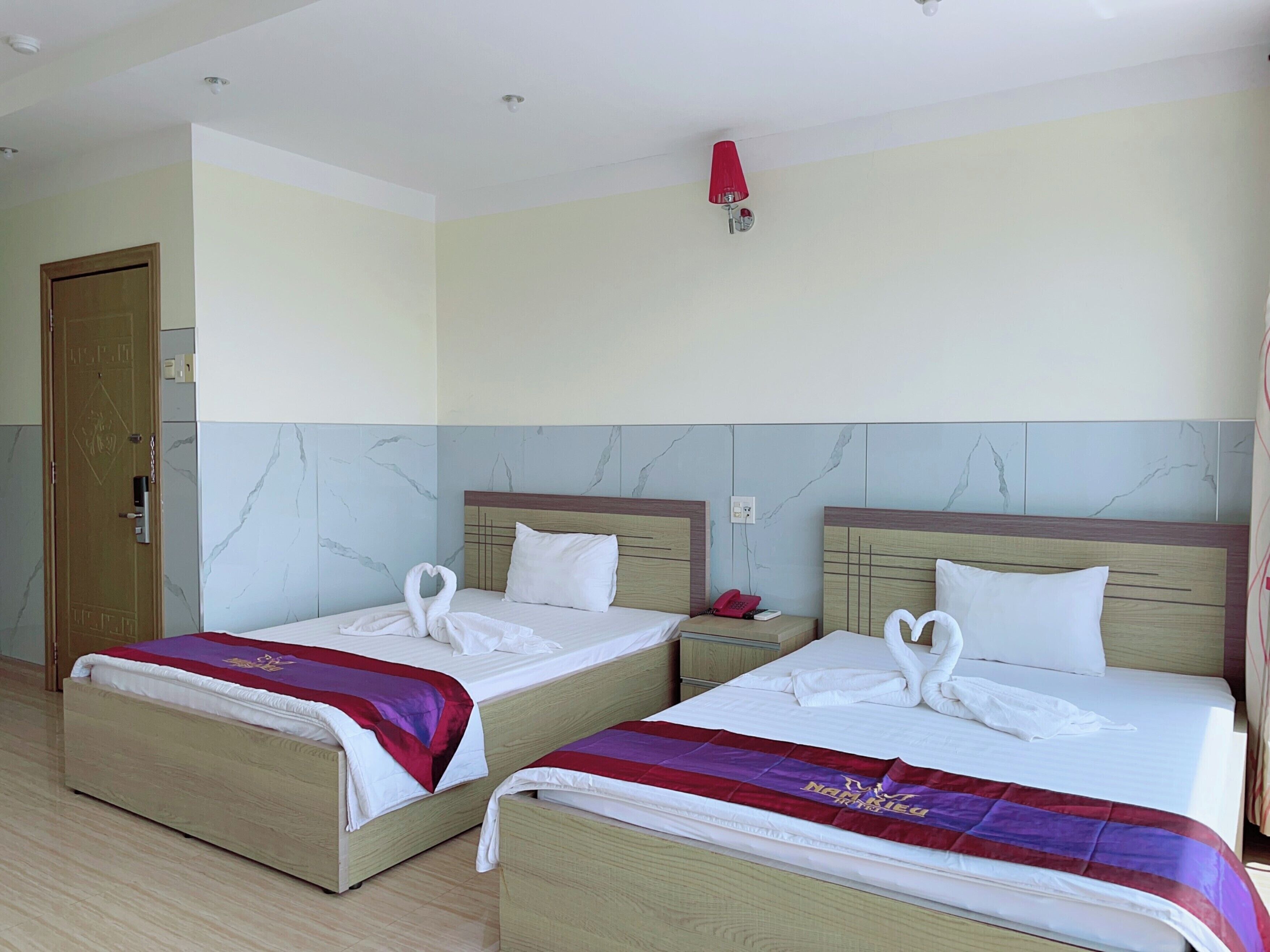 Une chambre d’hôtel avec deux lits, chacun orné d’une serviette en forme de cœur posée sur l’oreiller, d’une lampe rouge et d’une porte en bois.