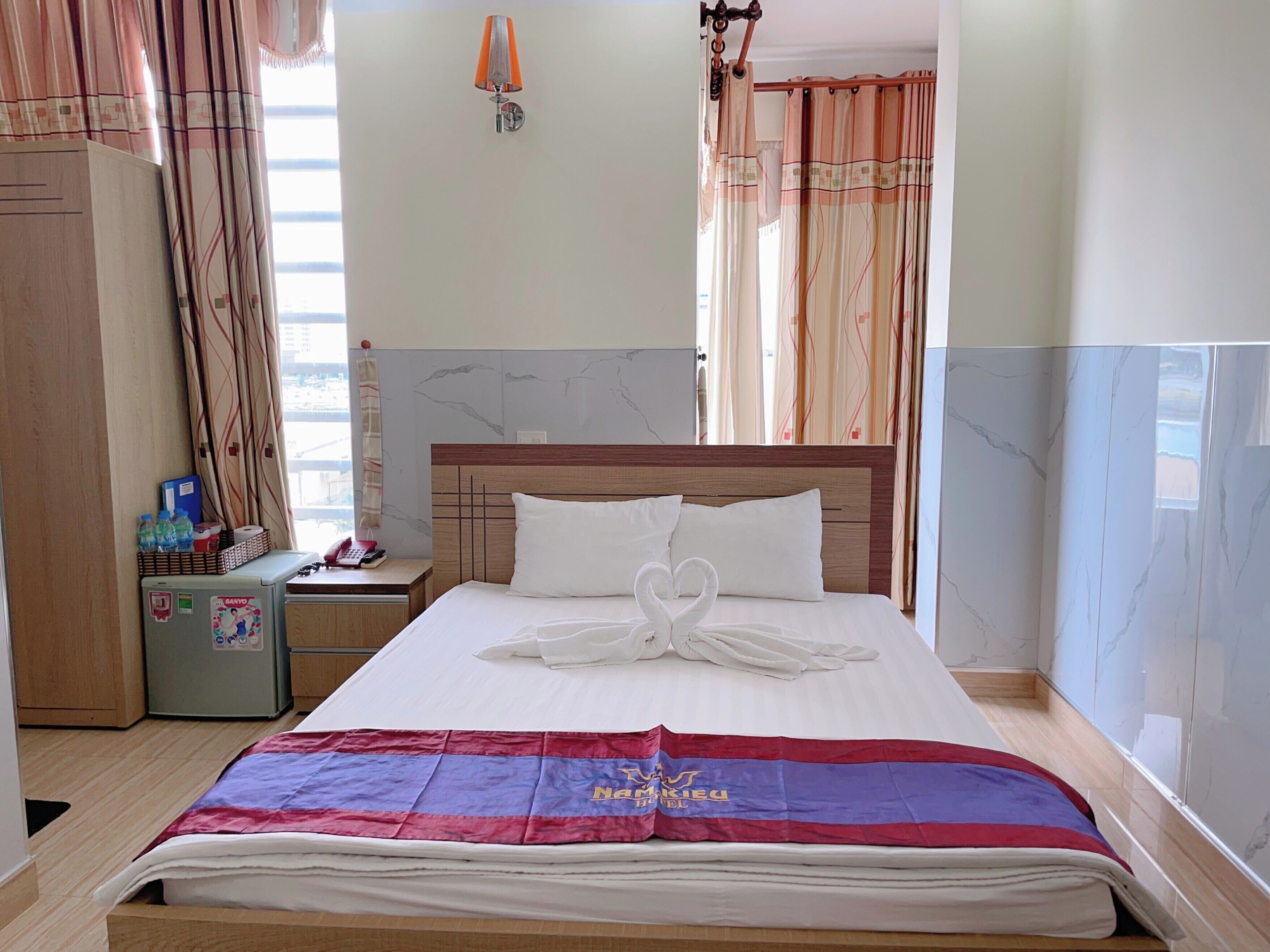 A hotel room with a neatly made bed, a nightstand with a lamp, a refrigerator, and a window with curtains.
