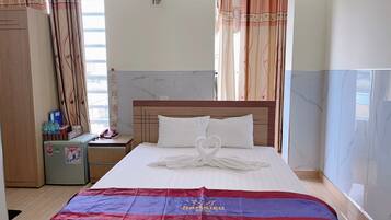 A hotel room with a neatly made bed, a nightstand with a lamp, a refrigerator, and a window with curtains.