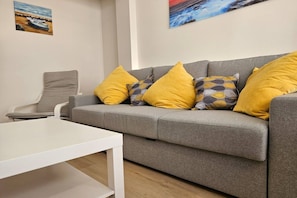 Un sofá gris con cojines amarillos y estampados, una mesa blanca y una silla.