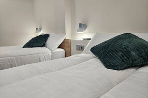 Deux lits simples avec du linge de lit blanc et des oreillers verts, une table de chevet avec une lampe et un luminaire fixé au mur.