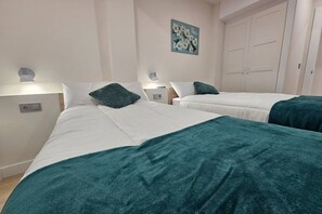Habitación de hotel con dos camas, cada una con ropa blanca de cama y almohadones color turquesa. Hay una obra de arte colgada en la pared sobre las camas y un armario con dos puertas a la derecha.