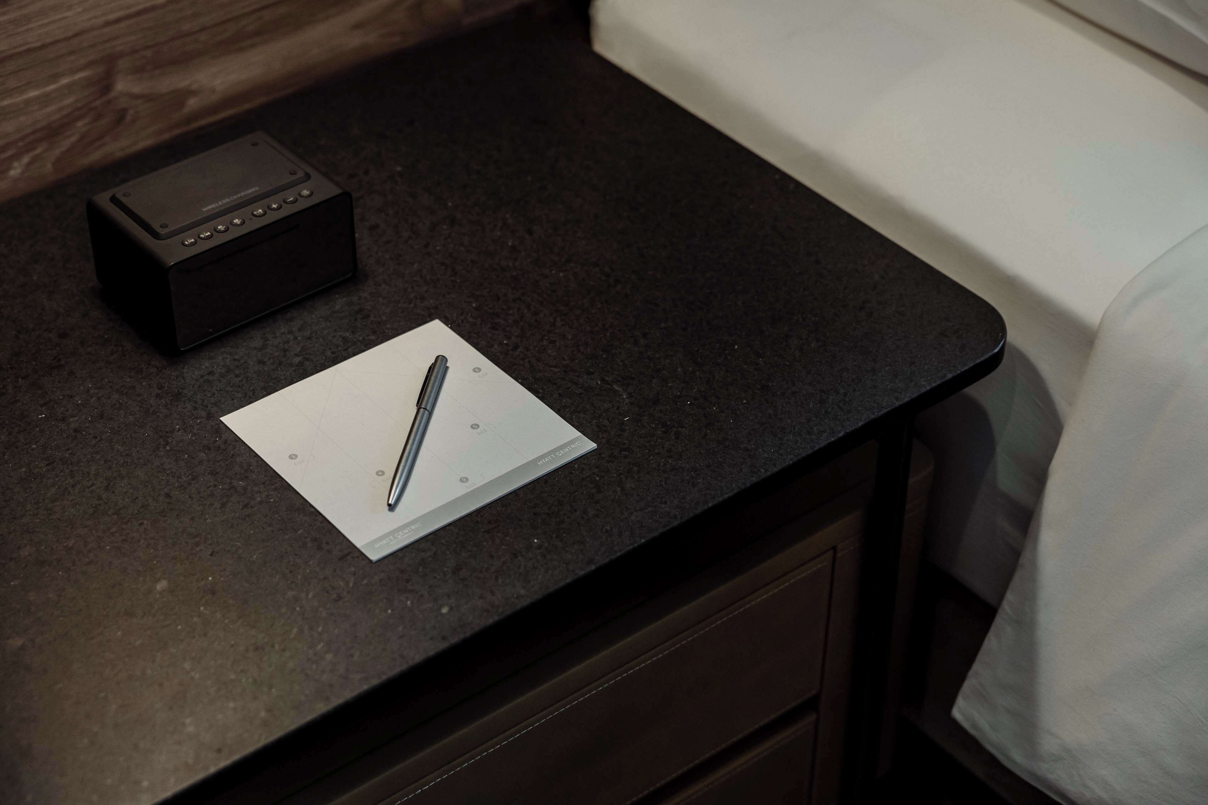 A small black electronic device on a dark countertop, a white piece of paper with a pen, and a white bed in the background.
