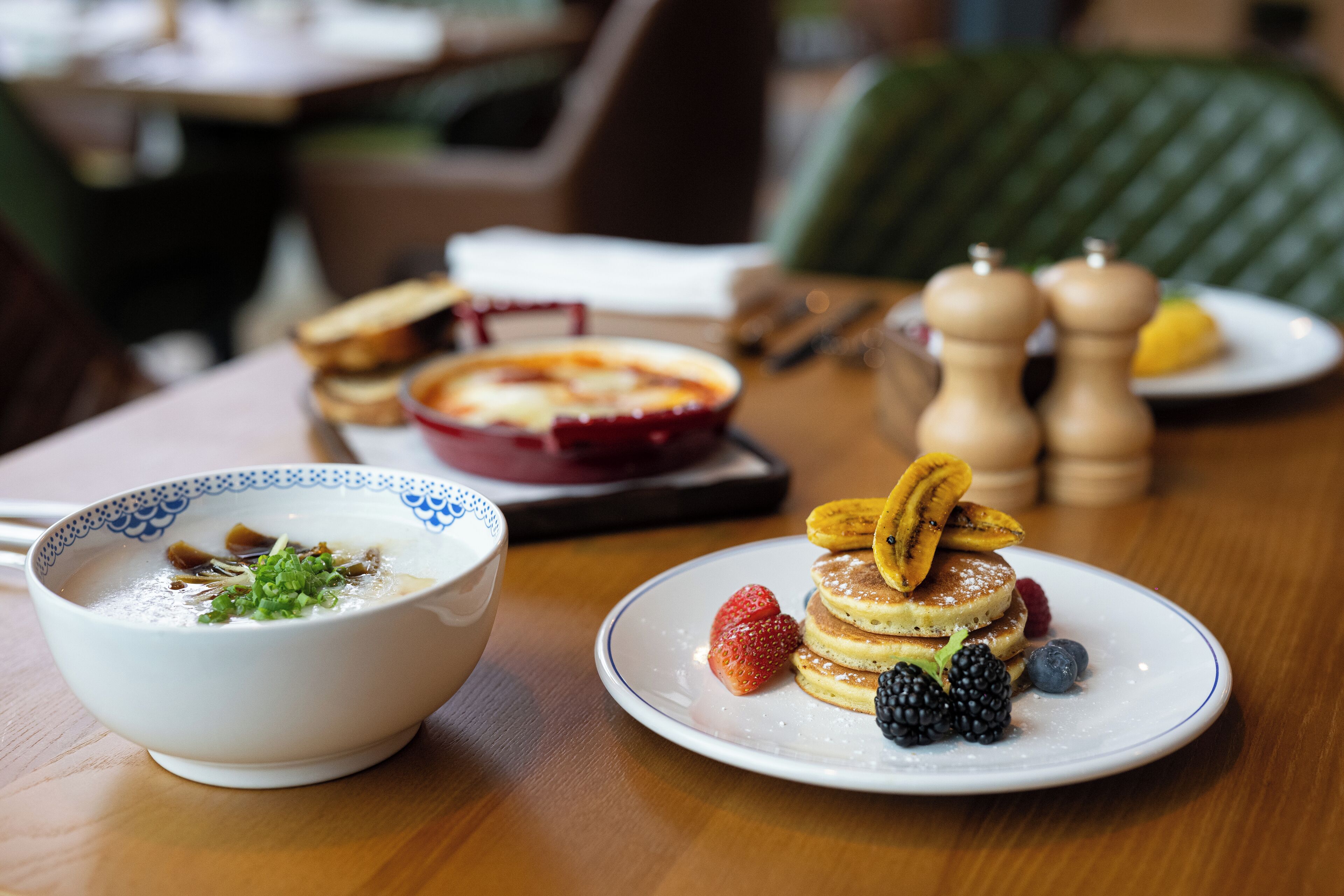 A dining table with a bowl of soup, a plate of pancakes with fruit, and a red dish in the background.