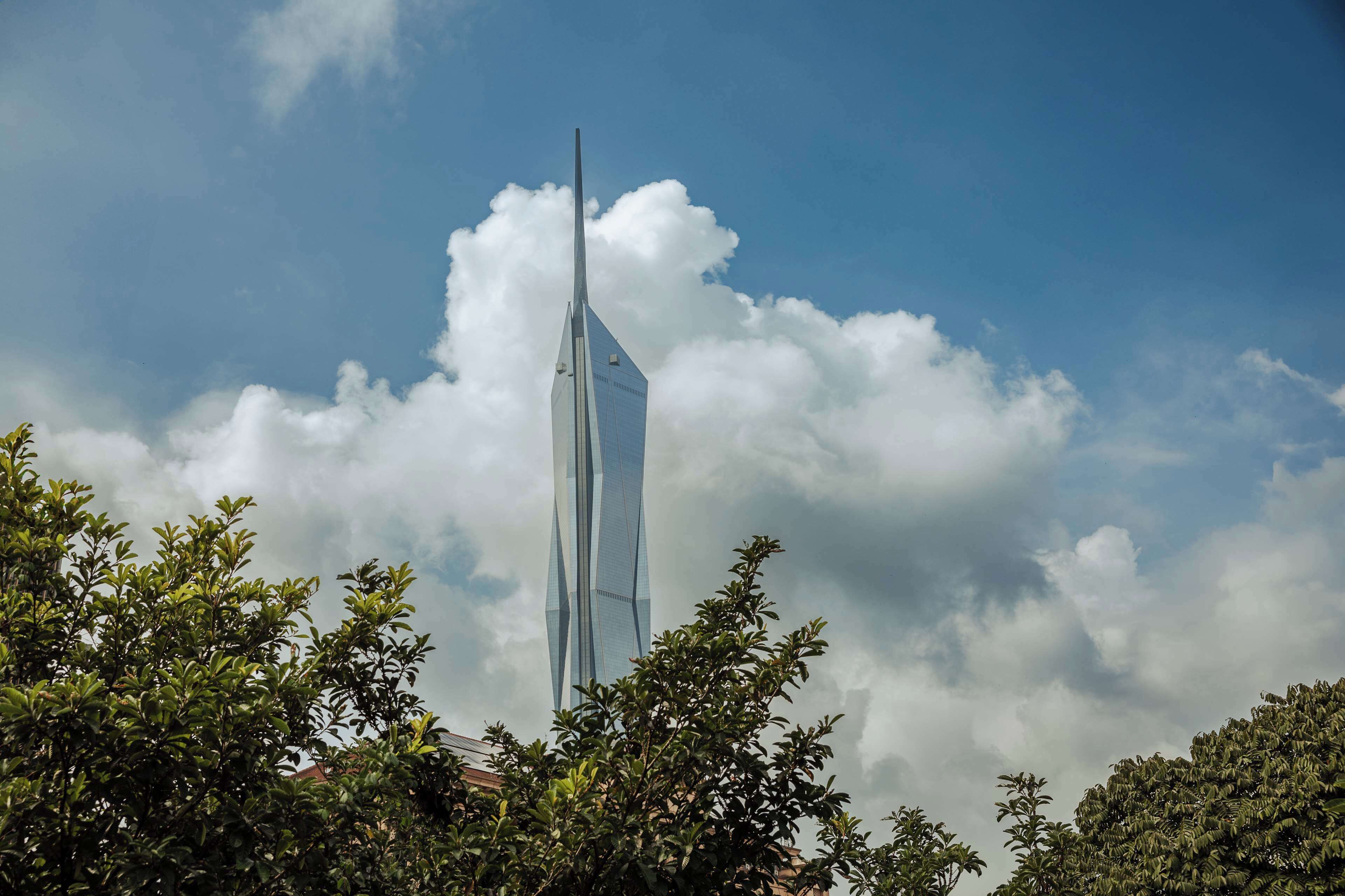 A tall, modern skyscraper with a pointed top, surrounded by dense foliage and partly cloudy sky.