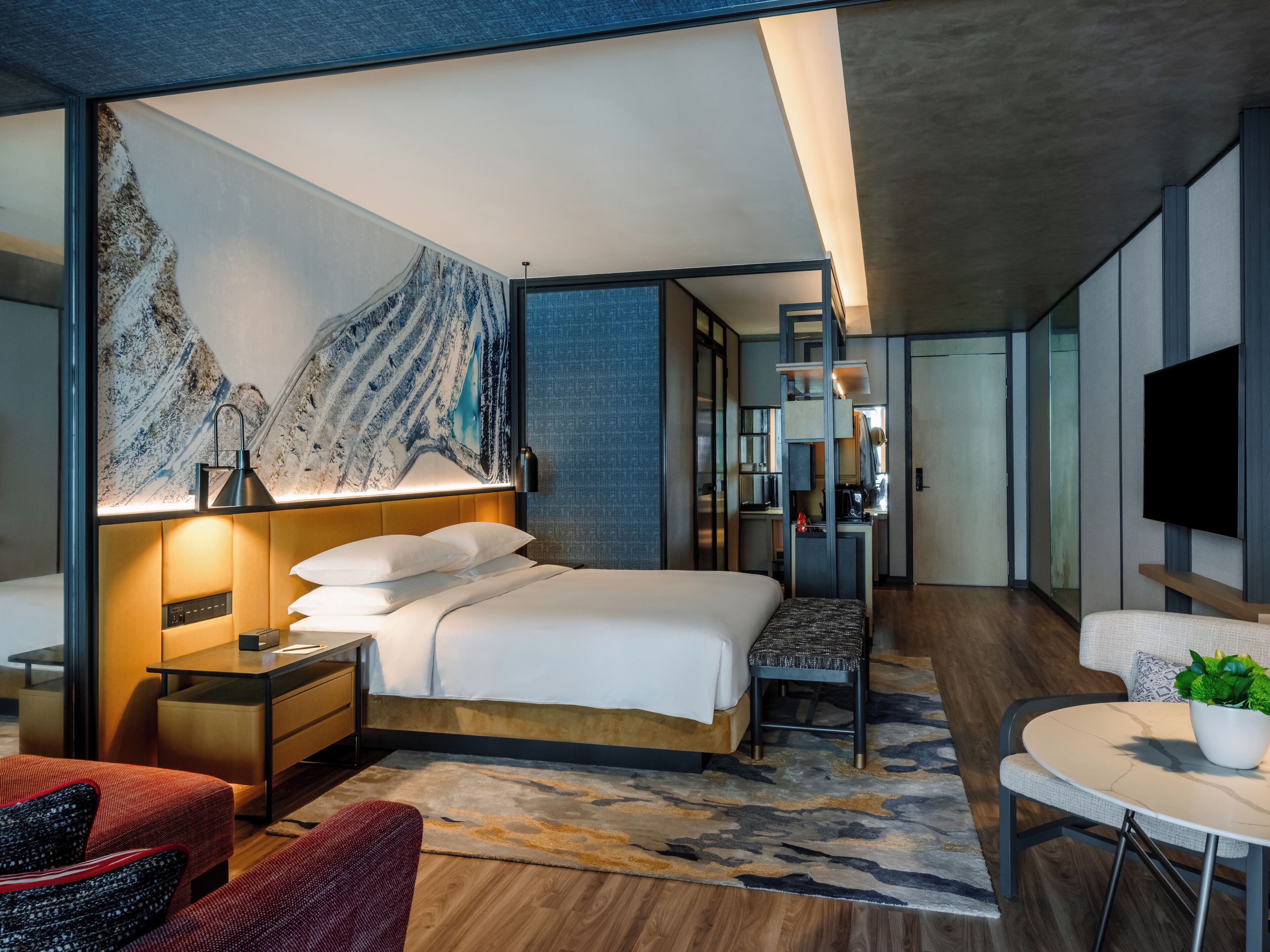 A modern hotel room with a large bed, a desk, a chair, a TV, and a painting on the wall.