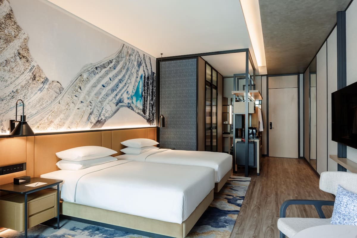 A modern hotel room with two beds, a desk, a flat-screen TV, and a large abstract painting on the wall.