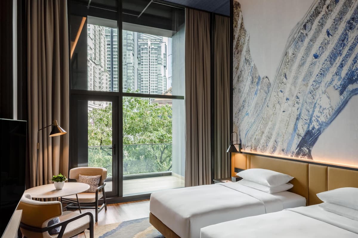 A hotel room with two beds, a dining area with a table and chairs, a large window with curtains, and a painting on the wall.