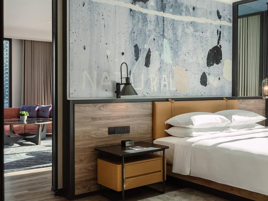 A modern hotel room with a large bed, a nightstand, a desk, and a view of the cityscape.