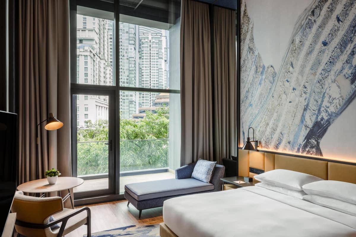 A modern hotel room with a large window, a bed, a sofa, a small table with a plant, and a painting on the wall.