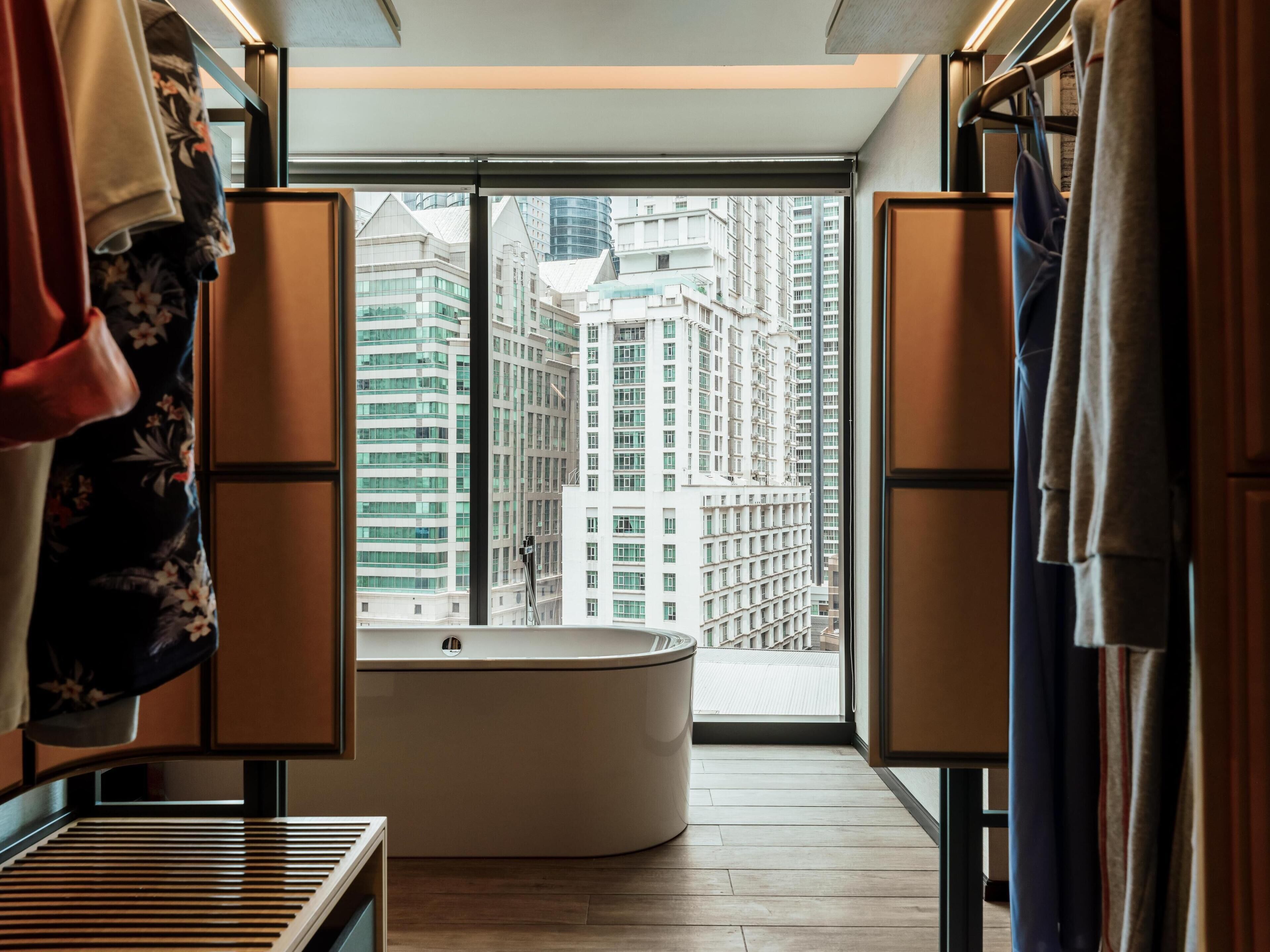 Un baño moderno con una bañera independiente, suelo de madera y vistas a edificios altos a través de amplios ventanales.