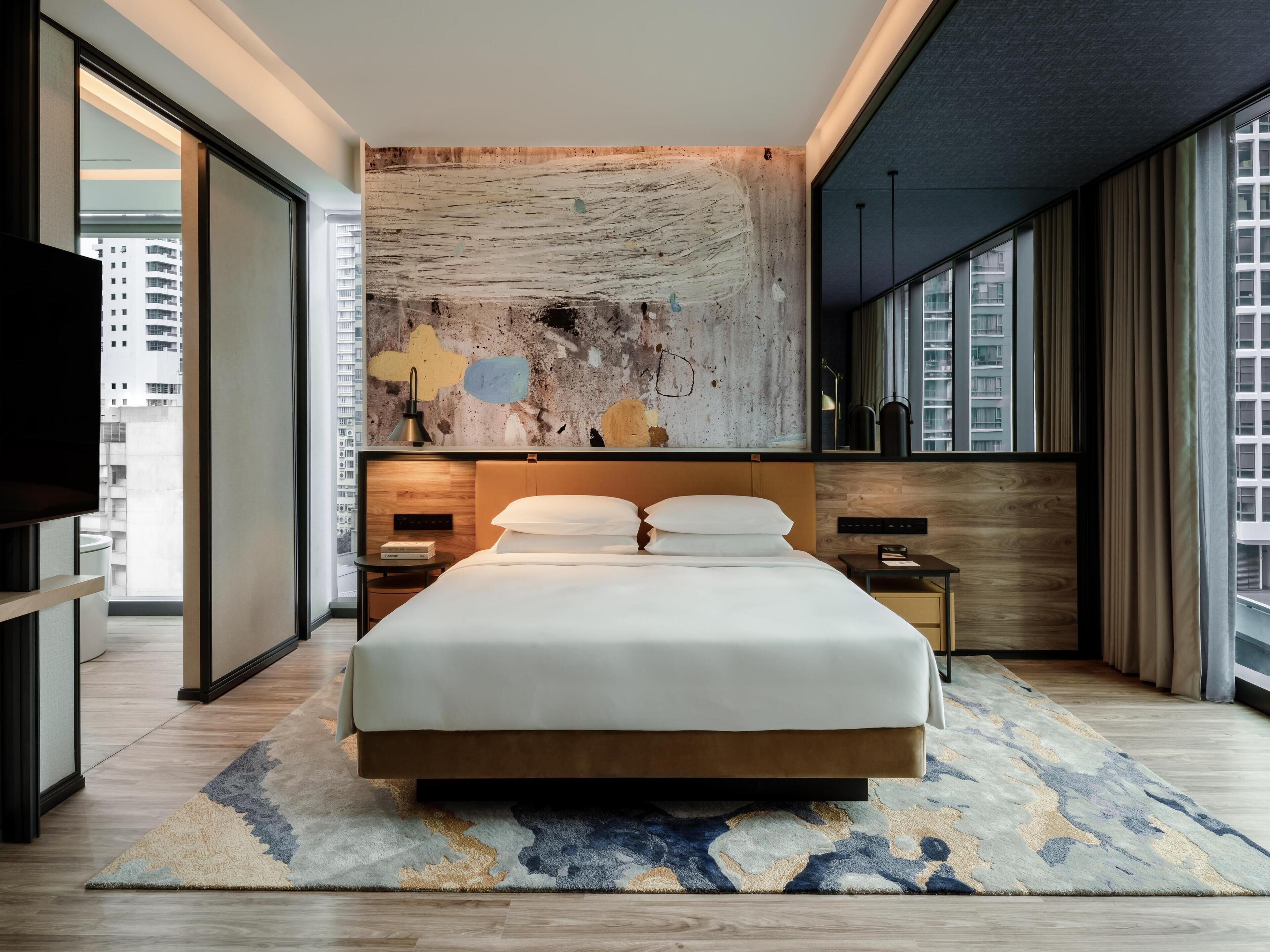 A modern hotel room with a large bed, a city view, a textured wall art, and a large window with curtains.